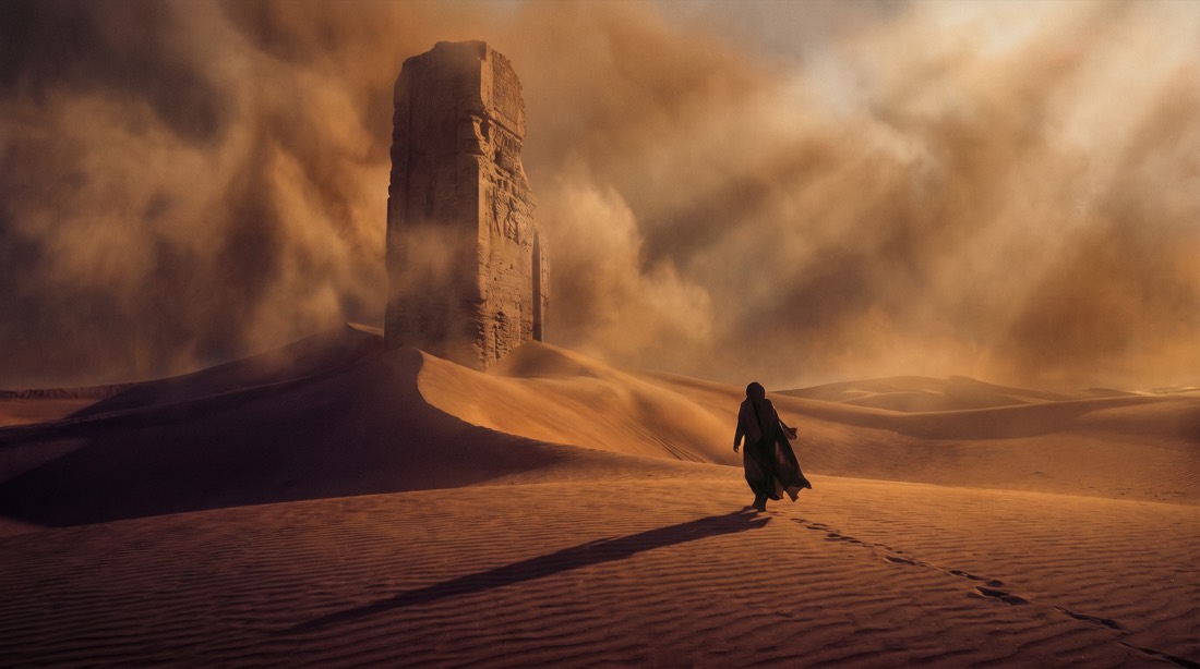 Vast desert at golden hour, a colossal weathered stone monolith half-buried in dunes, lone figure in flowing robes walking toward it casting a long shadow, wind whipping sand into the air, god rays piercing dust clouds, epic wide establishing shot, warm amber and deep shadow palette, Denis Villeneuve aesthetic, ultra-detailed, 70mm film grain. 16:9 aspect ratio